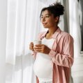 A pregnant woman holds a coffee mug and looks through sheer curtains out a window. 